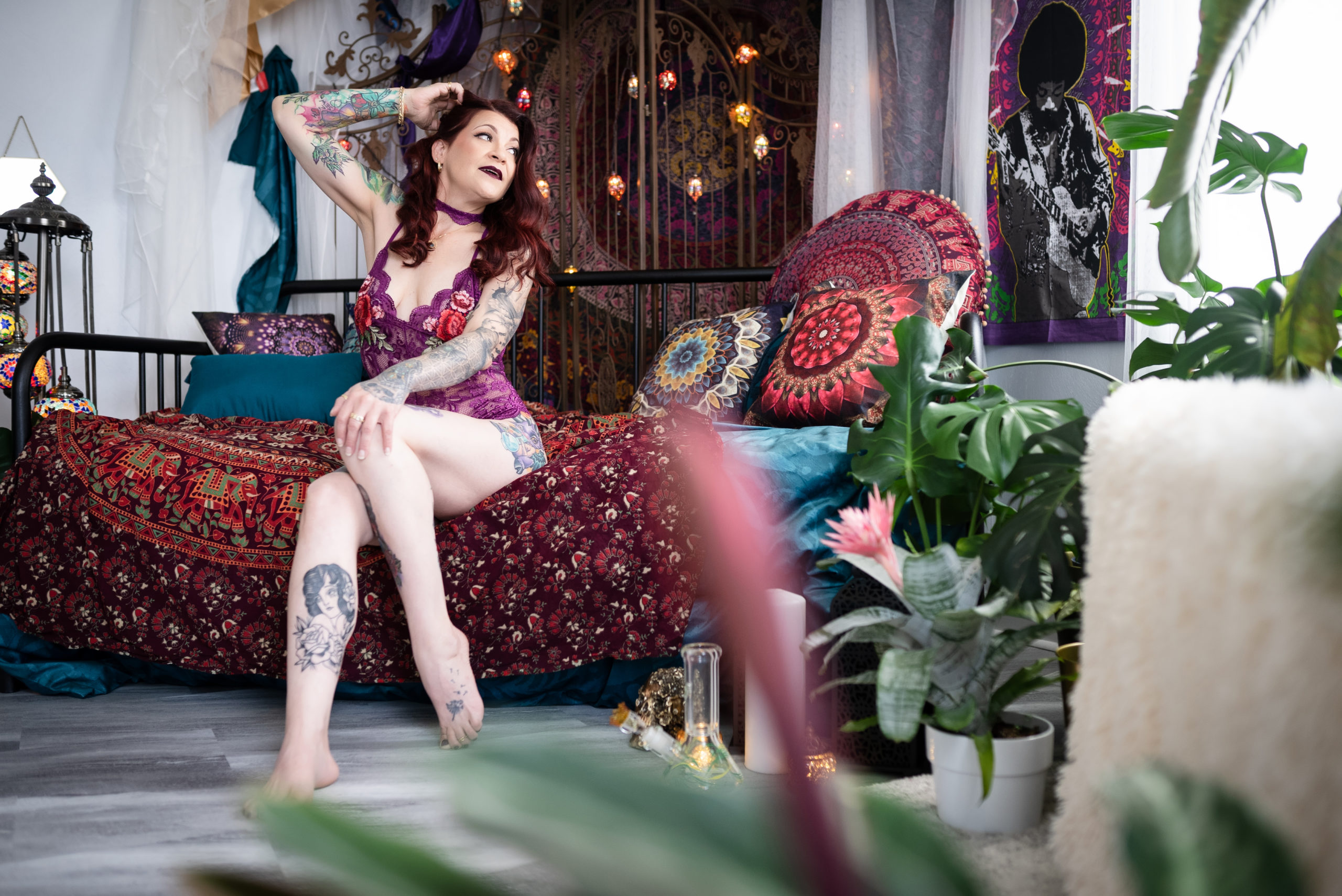 A woman with red hair and tattoos is wearing maroon lingerie while sitting on a bed during a boudoir photoshoot