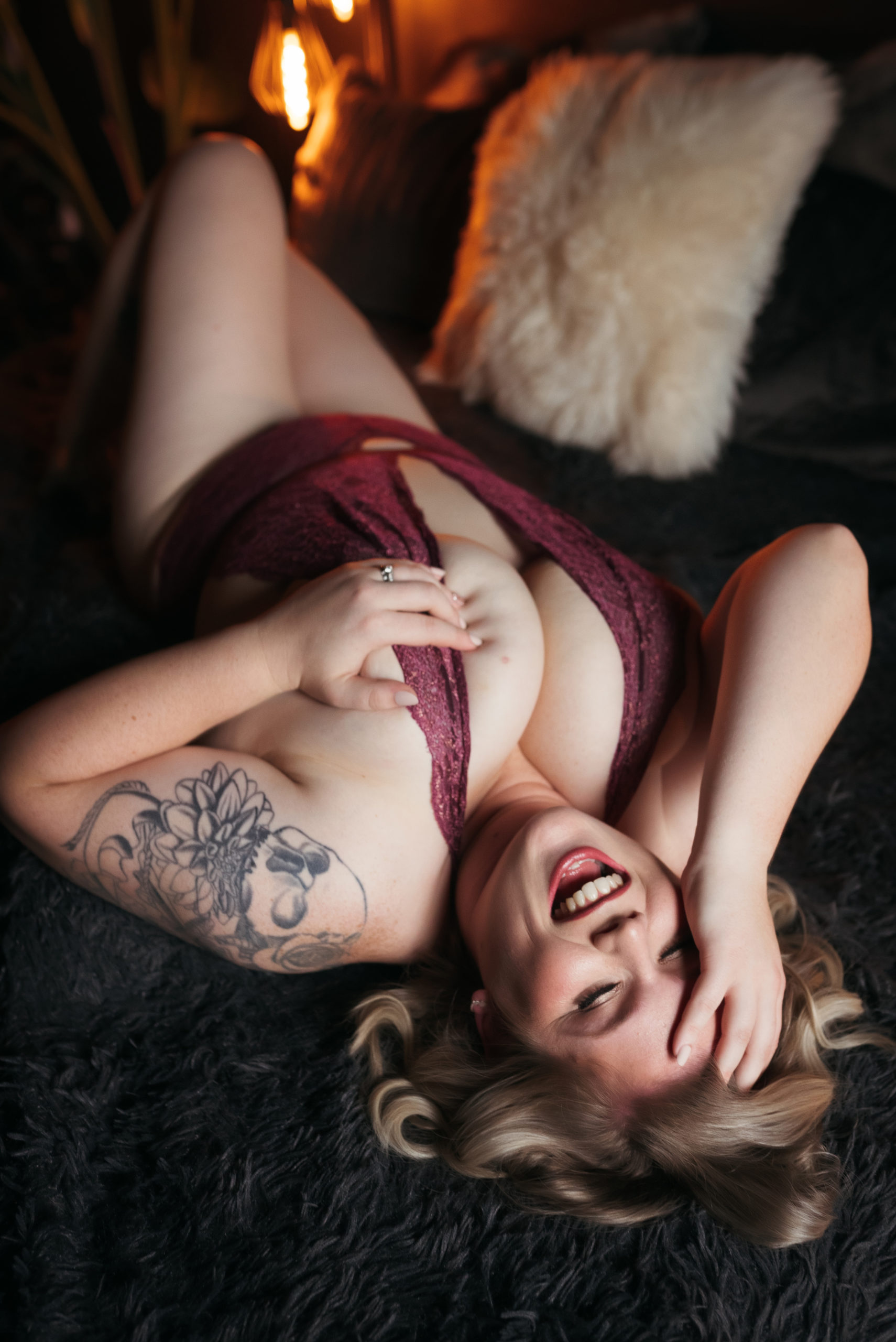 A blonde woman lies on a bed while laughing during a sensual boudoir photoshoot