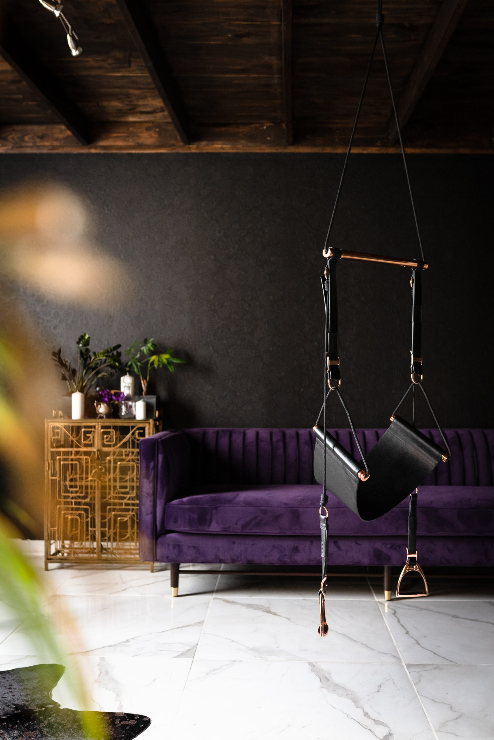 A Black leather sex swing hangs from the ceiling in front of a plush purple couch