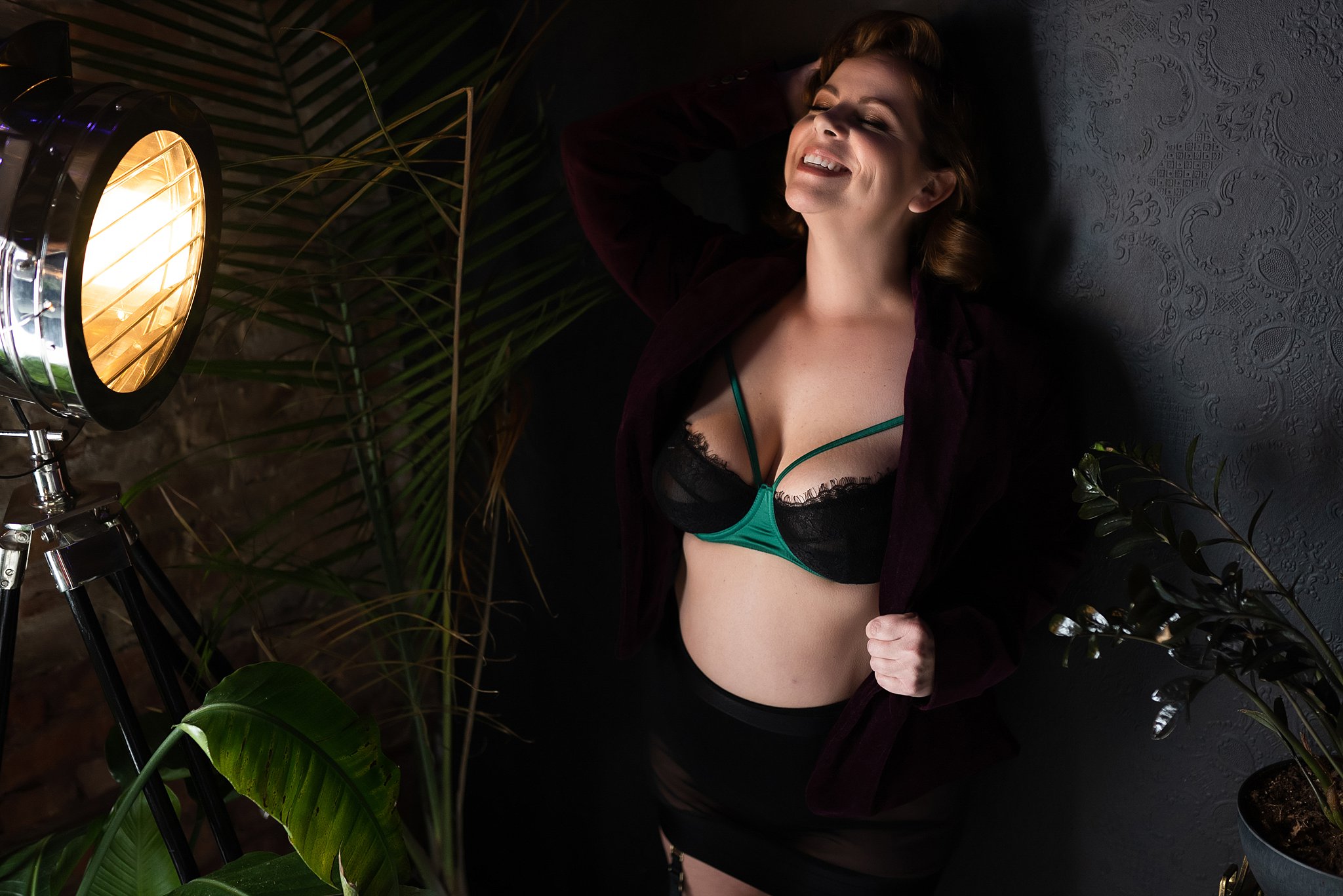 Miss P stands against a black damask textured wall during a 420 photoshoot with LeZandra Photography. She is facing a vintage studio light fixture while her right arm is raised and she runs her fingers through her hair. Miss P wears an emerald green satin bra with black lace and emerald green straps. Over her bra she is wearing a burgundy velvet jacket with black high waisted panties and a black sheer skirt.