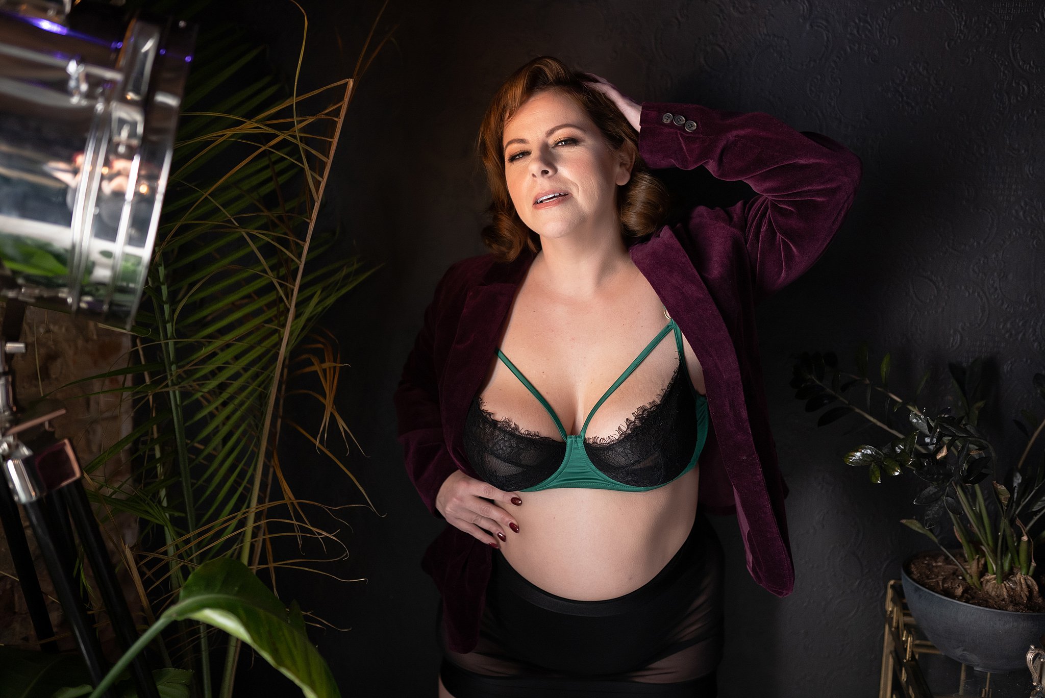 Miss P stands against a black damask textured wall during a 420 photoshoot with LeZandra Photography. She is happy and laughing. Miss P wears an emerald green satin bra with black lace and emerald green straps. Over her bra she is wearing a burgundy velvet jacket with black high waisted panties and a black sheer skirt.