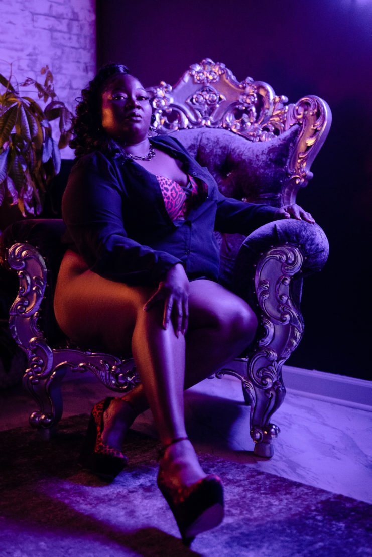 Miss D sits on a plush purple couch during a boudoir session with LeZandra Photography in Norfolk Virginia Boudoir Studio surrounded by purple neon lights